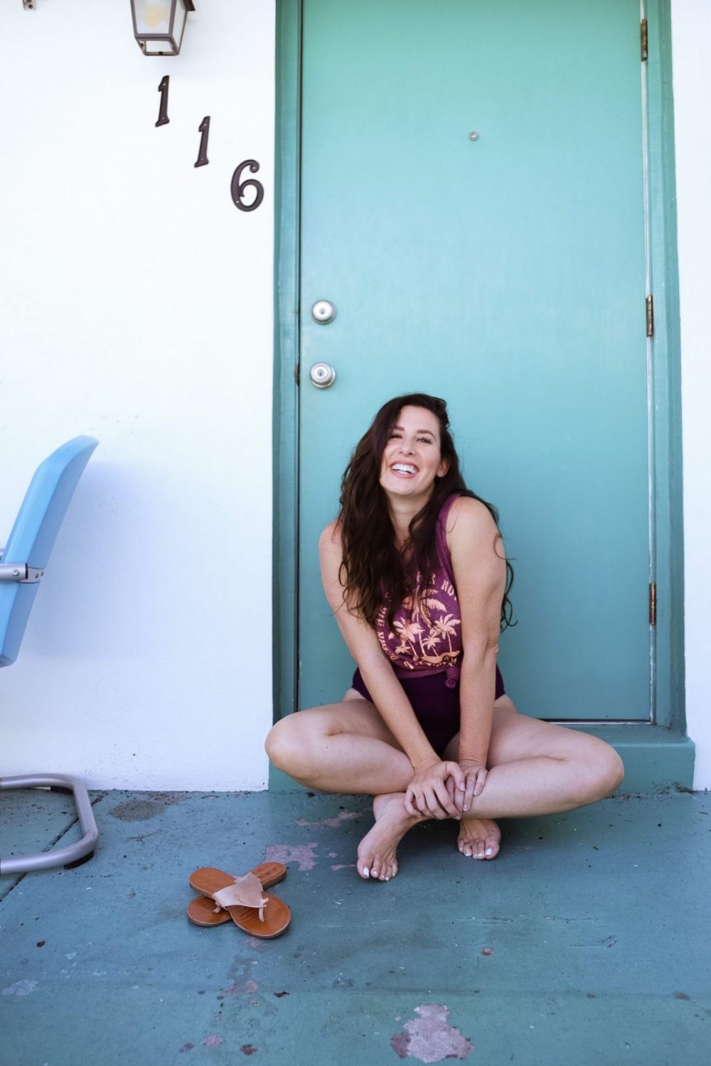 Style on a Budget: 9 Best Swimsuits for Moms |Swimsuits for Moms by popular Florida fashion blog, Fresh Mommy Blog: image of a woman sitting outside her Palm Court motel room and wearing a purple tank top with a pink palm tree print over a black one piece swimsuit. 